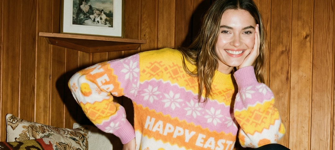 Smilende kvinde i lyserød og gul sweater med Happy Easter-motiv foran træpanelvæg